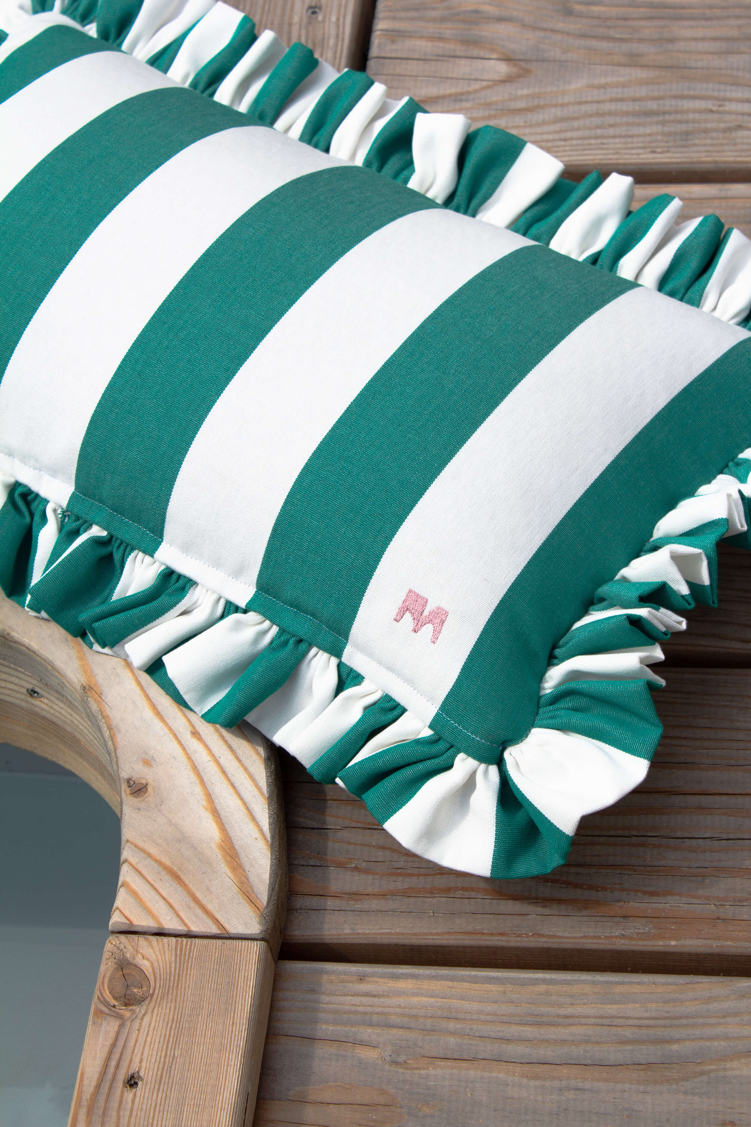 Grün-weiß gestreiftes Outdoor-Kissen mit Rüschenkante von another mondaen auf Holzoberfläche – dekoratives Designer-Kissen für Terrasse, Balkon und Garten.